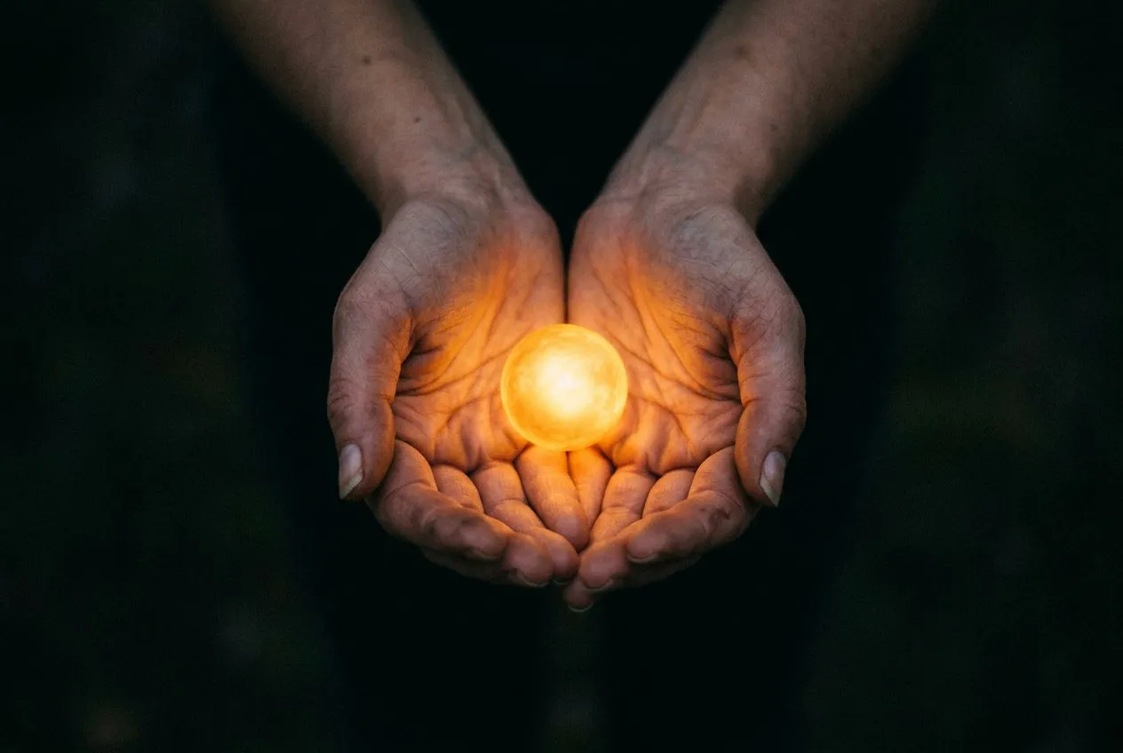 雙手捧住一顆發光的小光球,從略高角度拍攝,雙手位於畫面下方居中,周圍黑暗向邊緣消退,溫暖金光映照在皮膚上